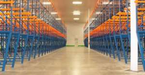 Vista interior de un almacén con estanterías metálicas azules y naranjas dispuestas en filas, creando un pasillo amplio y despejado con un suelo de concreto pulido