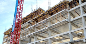 Detalle de un edificio en construcción, mostrando la estructura de hormigón y andamios, con una grúa roja al lado y un cielo despejado