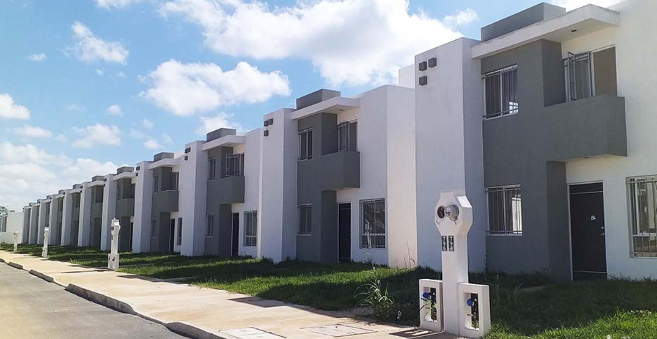 Vista de un conjunto de casas modernas con fachadas en blanco y gris, alineadas a lo largo de una calle, con jardines frontales y un cielo azul con nubes