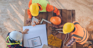 Cuatro trabajadores de la construcción revisando planos y datos en una computadora portátil, rodeados de herramientas y equipo de seguridad, sobre una mesa de madera en el sitio de construcción
