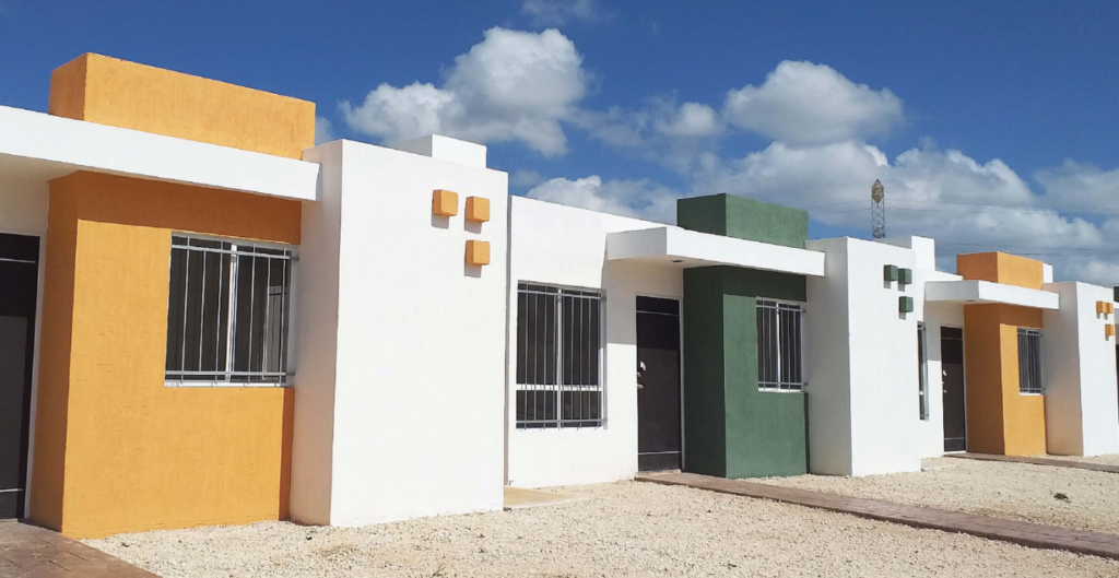 Vista de un conjunto de casas modernas, con fachadas en colores naranja, blanco y verde, y ventanas con rejas, sobre un área de grava, bajo un cielo azul con nubes