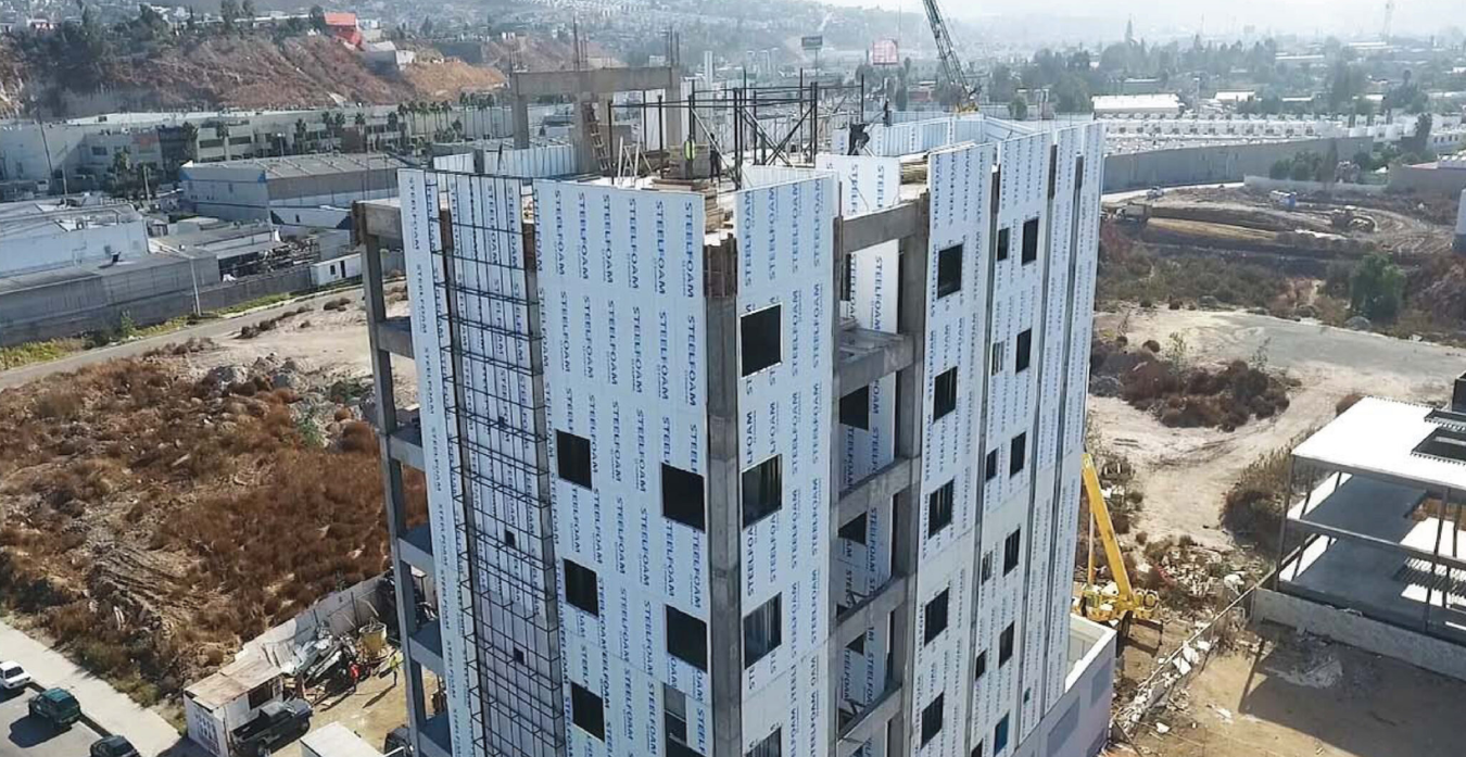 Vista aérea de un edificio en construcción de varios pisos, con paneles exteriores colocados de steelfoam de fanosa, grúas y maquinaria en el sitio, en un área desértica alrededor, bajo un cielo despejado