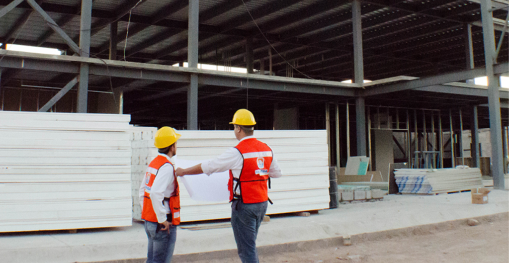 Dos ingenieros con cascos y chalecos de seguridad revisan planos en una obra en construcción con estructuras de acero y materiales de construcción apilados en el fondo