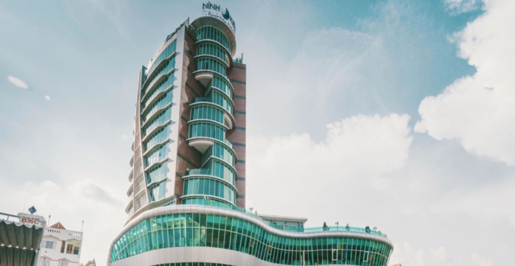 Vista desde abajo de un edificio moderno con diseño curvo y grandes ventanales, resaltando su estructura elegante y contemporánea bajo un cielo claro y nubes esponjosas