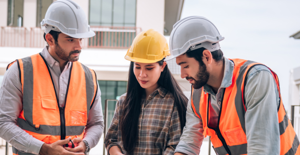 Tres profesionales en un sitio de construcción, dos hombres con cascos y chalecos reflectantes, discutiendo planos con una mujer que lleva un casco amarillo, todos mirando hacia un documento sobre una mesa