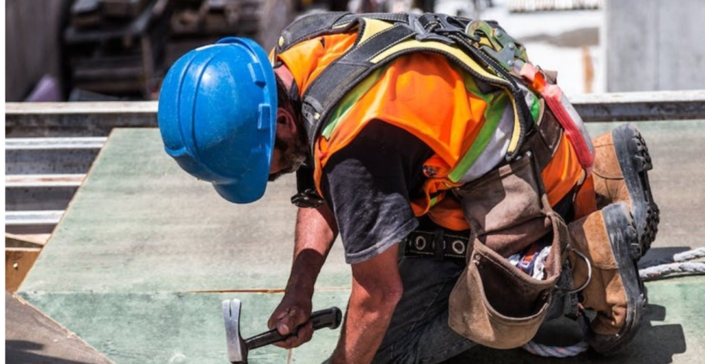 Un trabajador de la construcción arrodillado en una plataforma, usando un casco azul y chaleco reflectante, mientras clava un clavo en una tabla de madera, rodeado de materiales de construcción