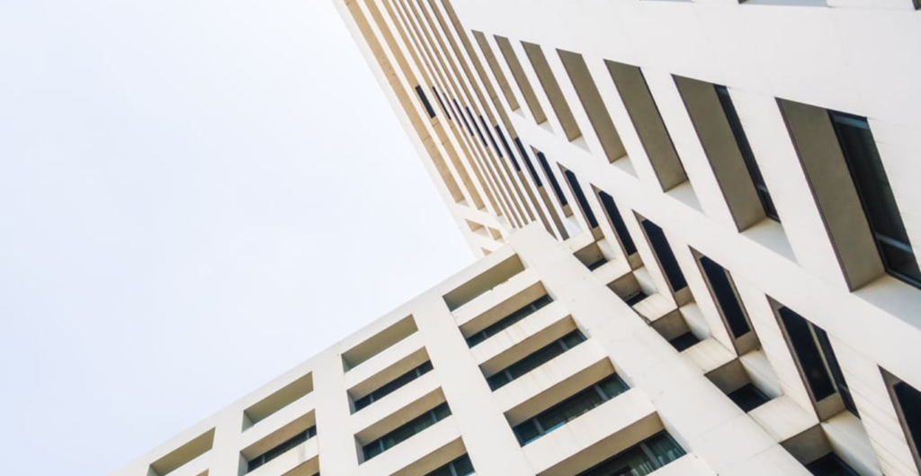 Vista desde abajo de un edificio moderno, mostrando una fachada blanca con ventanas alargadas y líneas arquitectónicas que se elevan hacia el cielo, bajo un fondo nublado