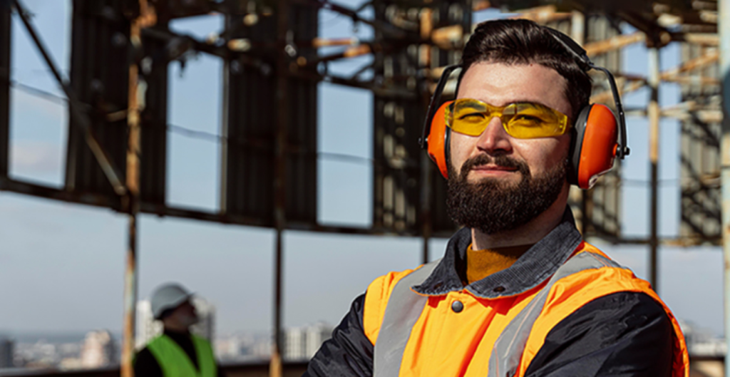 Un trabajador de la construcción con barba, usando gafas de seguridad y orejeras, luciendo un chaleco reflectante mientras sonríe, con una estructura metálica de fondo y otro trabajador parcialmente visible