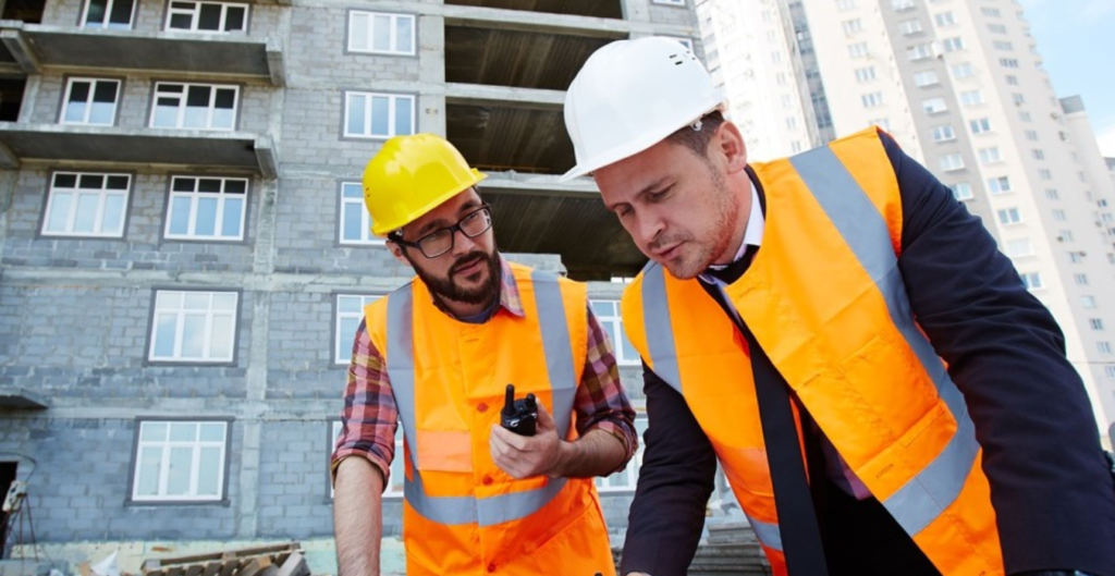 Dos hombres en un sitio de construcción, uno con chaleco reflectante y casco amarillo, el otro con traje y casco blanco, revisando planos sobre una mesa, con un edificio en construcción de fondo