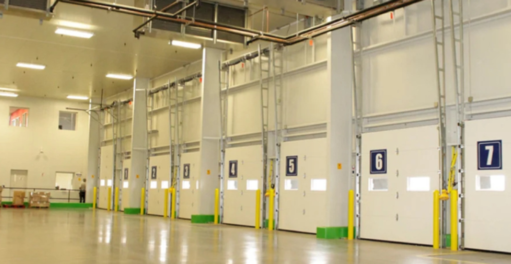 Interior de un almacén moderno con varias puertas de carga numeradas del 5 al 7, paredes claras y un suelo limpio, con estanterías visibles y un ambiente bien iluminado