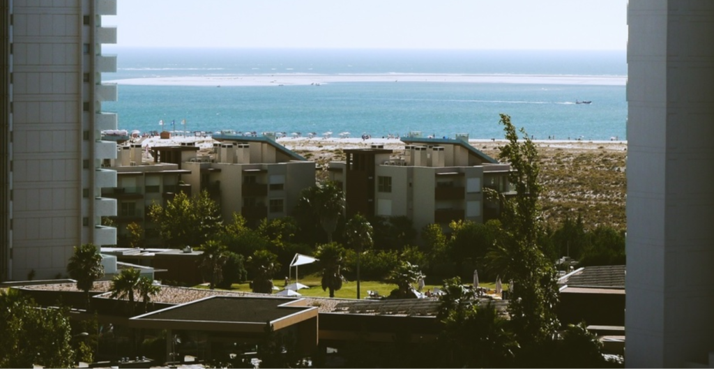 Vista panorámica de un paisaje costero, con edificios y áreas verdes en primer plano, y el mar azul y la playa en el fondo, bajo un cielo soleado