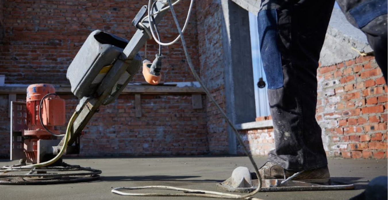 Vista cercana de un trabajador en una obra, utilizando una pulidora de superficie sobre un piso de cemento recién vertido, con paredes de ladrillo en el fondo