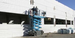 Dos trabajadores en una plataforma elevadora instalando paneles aislantes de EPS (Insulpanel) en el muro exterior de una nave industrial en construcción.