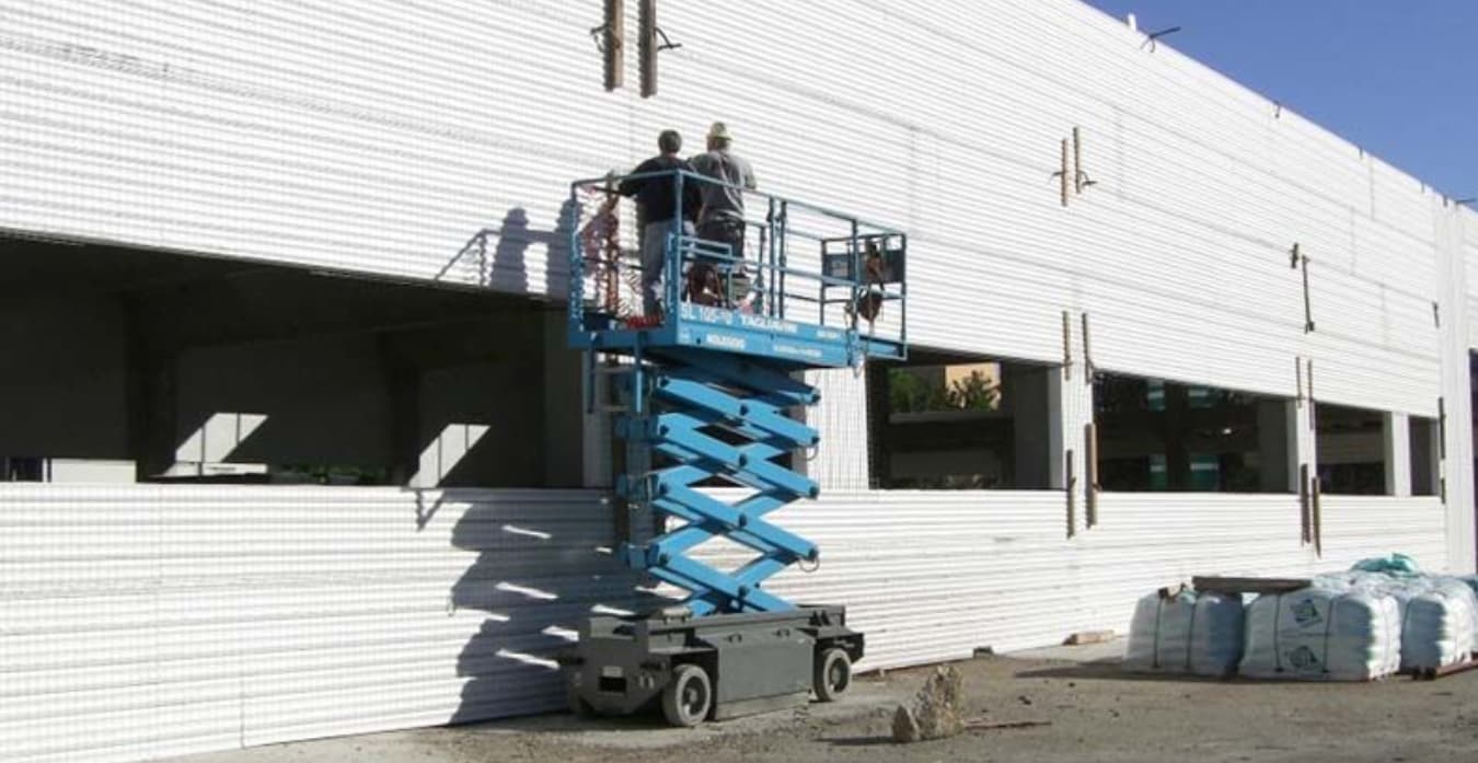 Dos trabajadores en una plataforma elevadora instalando paneles aislantes de EPS (Insulpanel) en el muro exterior de una nave industrial en construcción.