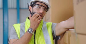 Primer plano de un trabajador de almacén o supervisor de logística, con barba, casco blanco y gafas de seguridad, hablando por un radio de comunicación (walkie-talkie) mientras se apoya en una pila de cajas de cartón.